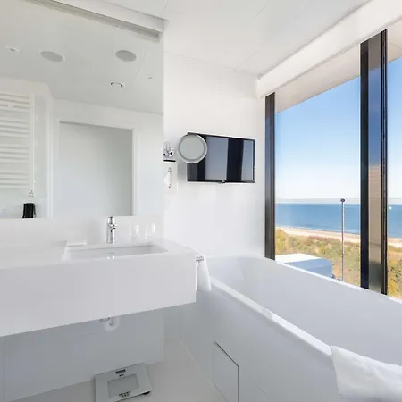 Radisson Blu Resort Swinoujscie Exterior photo The photo shows a modern bathroom with a minimalist design. It features a clean, white aesthetic with a large window offering a view of the beach and ocean. There's a bathtub positioned by the window and a sleek sink area with a mirror above it. A fl